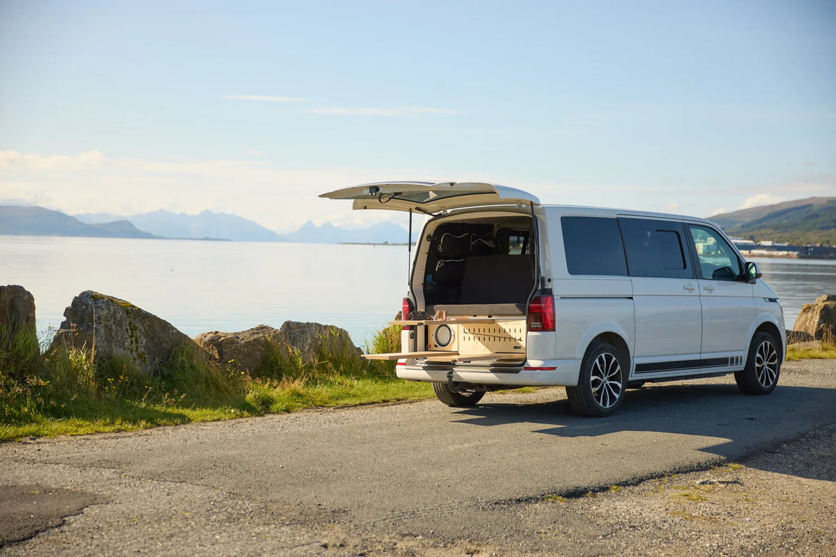 Silberner Campervan mit CampBoks und geöffnetem Heck an einem See mit Bergen.
