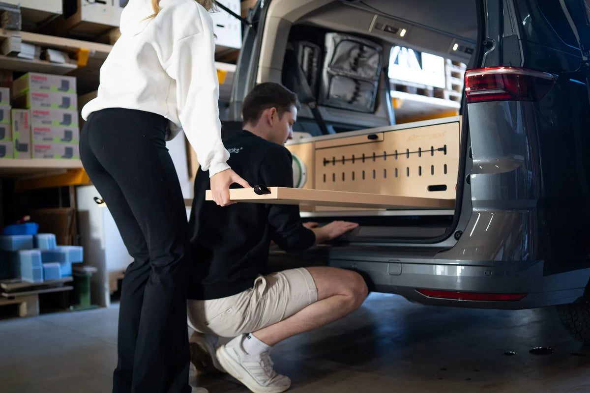 Zwei Personen setzen das CampBoks-Modul am geöffneten Fahrzeugheck in der Werkstatt ein.