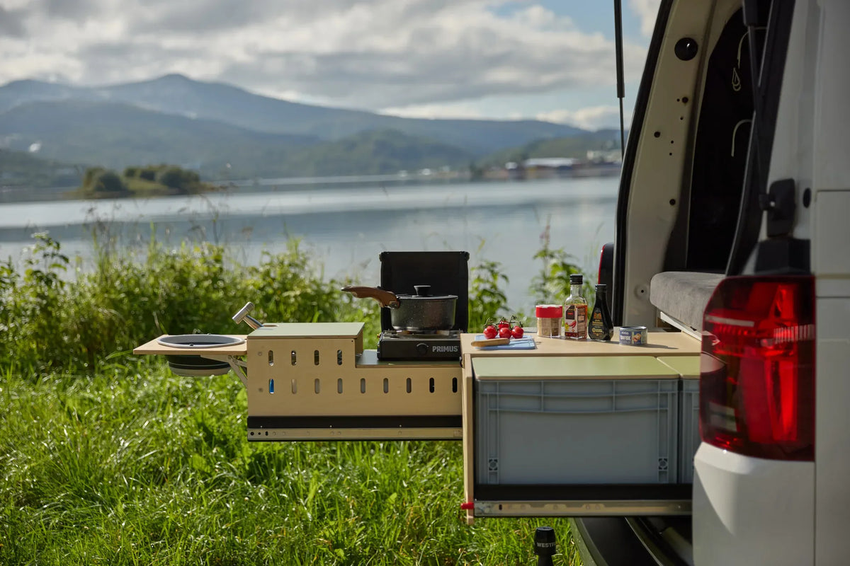 Ausgezogene CampBoks-Heckküche mit Gaskocher, Topf und Arbeitsfläche vor einem See.
