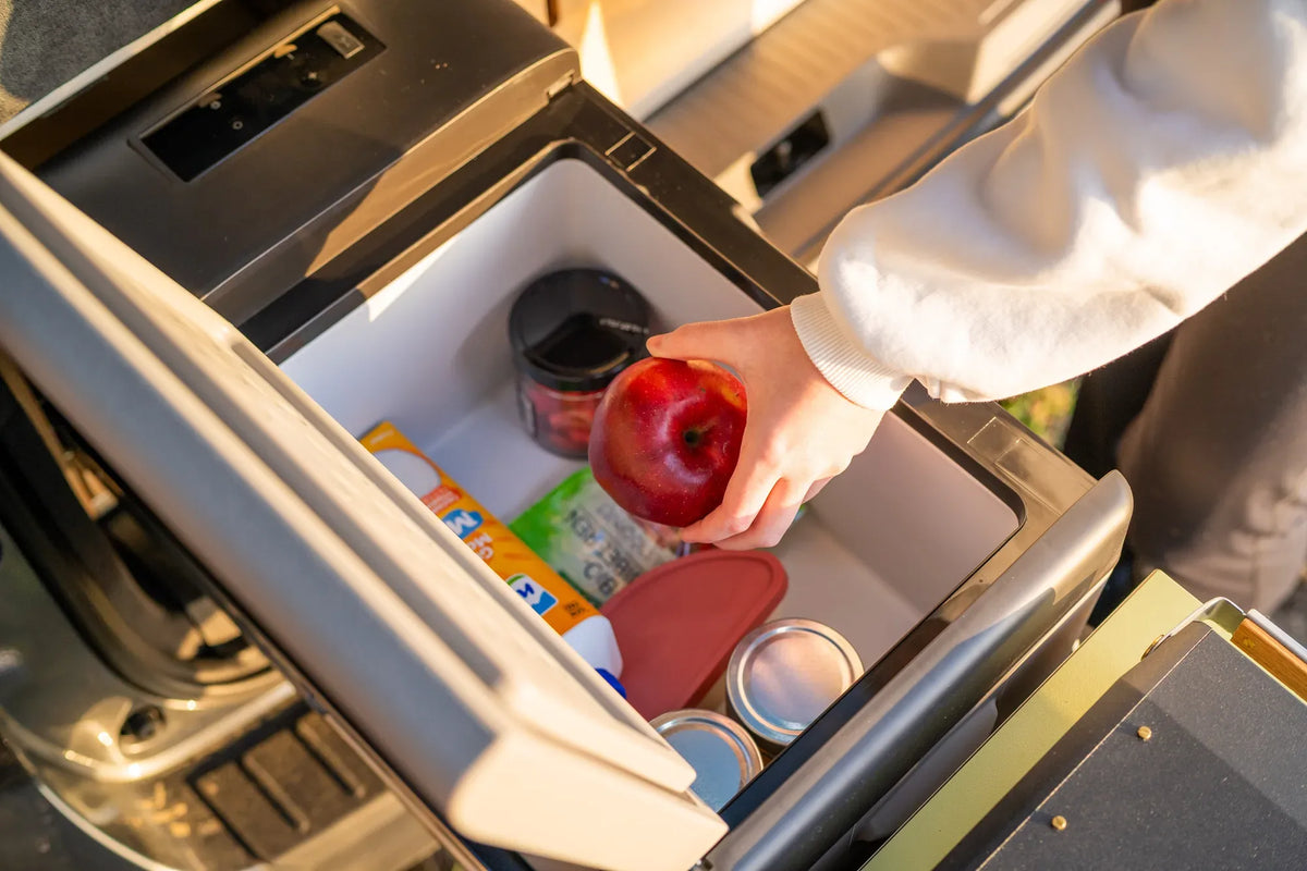 CampBoks Geöffnete Kompressor-Kühlbox mit Lebensmitteln; eine Hand legt einen roten Apfel hinein.