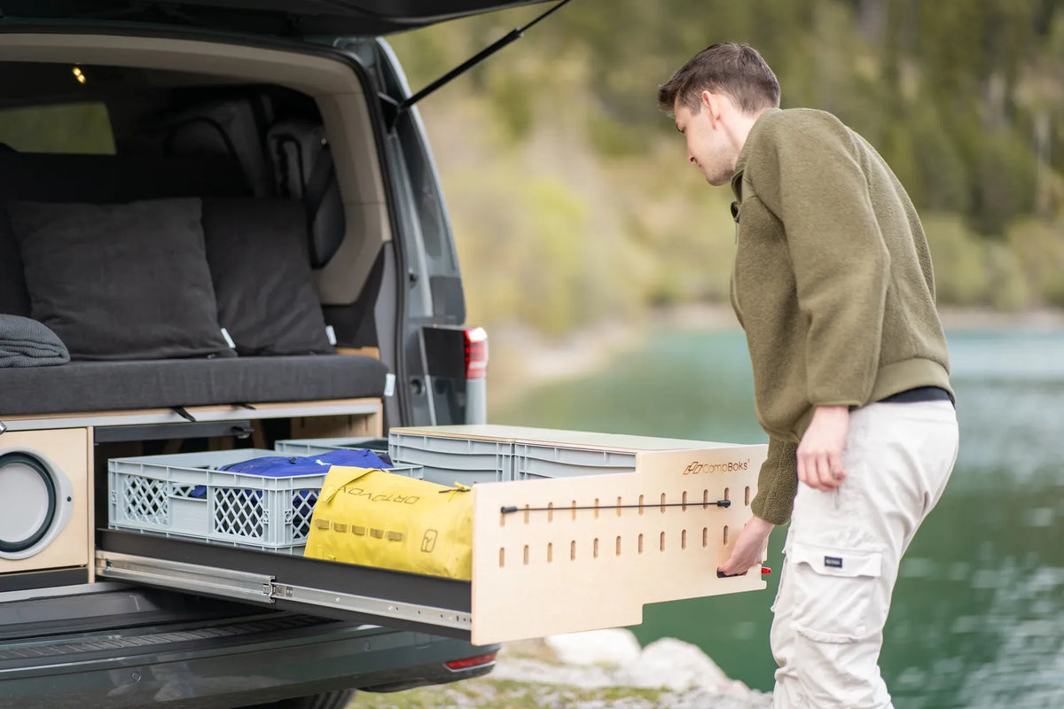 Ein Mann zieht das CampBoks-Stauraummodul mit Euroboxen und gelber Tasche am Fahrzeugheck heraus.