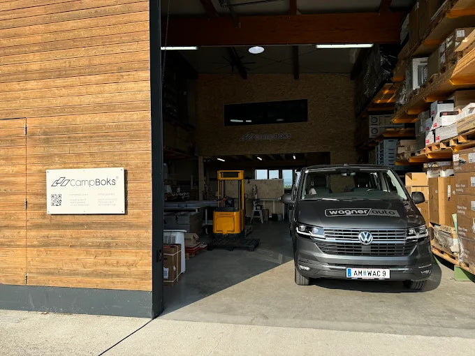 Halle mit Holzfassade und CampBoks-Schild, im offenen Tor steht ein VW-Bus